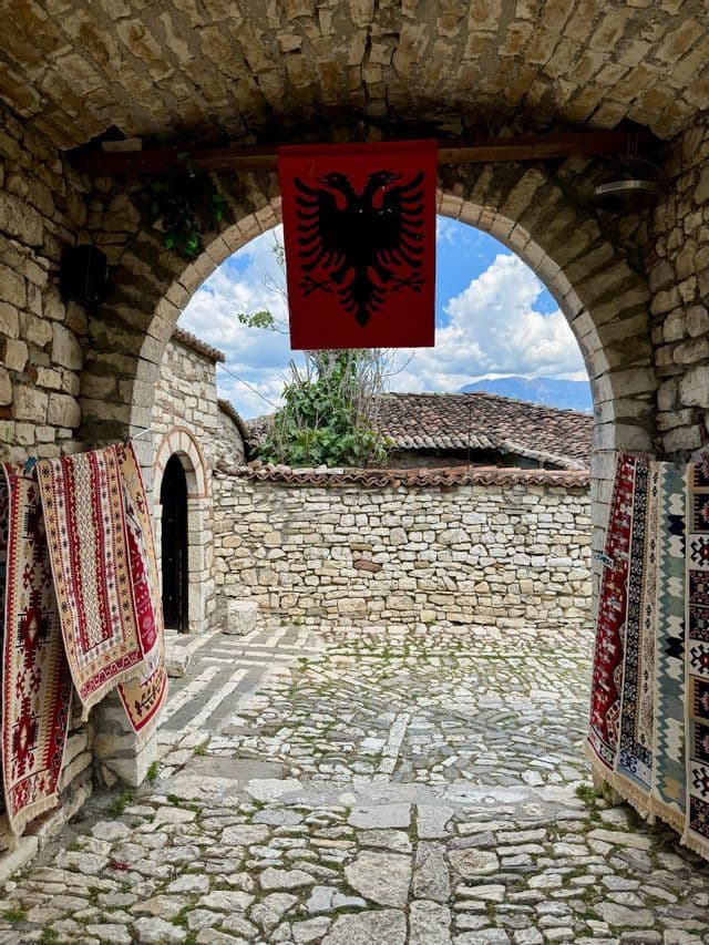 Una bandiera albanese e tappeti tradizionali pendono all'interno di un arco in pietra che conduce a un cortile acciottolato.