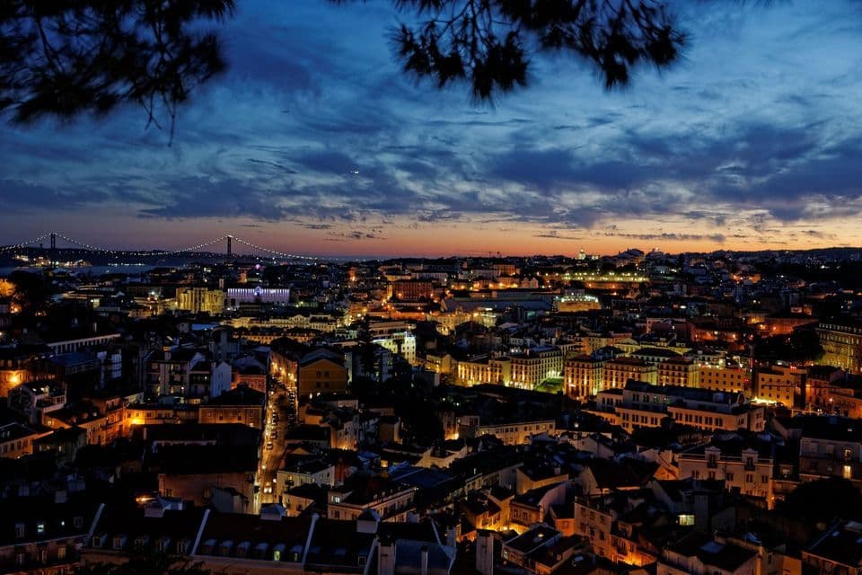 Vista panorámica de una extensa ciudad al atardecer, con edificios iluminados y un gran puente en el horizonte.