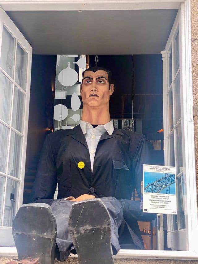 Una marioneta grande, de aspecto humano, con una chaqueta negra, sentada en el alféizar de una ventana abierta y con los pies en alto.