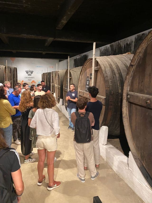 Un guía habla a un grupo de viaje de WeRoad durante un recorrido por una bodega con grandes barricas de vino de madera.