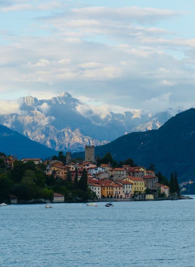 A colorful lakeside village with a stone tower is framed by large mountains under a partly cloudy sky.