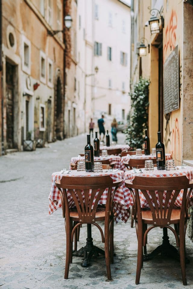 Empty restaurant tables with red and white checkered tablecloths and wine bottles set on a narrow cobblestone street.