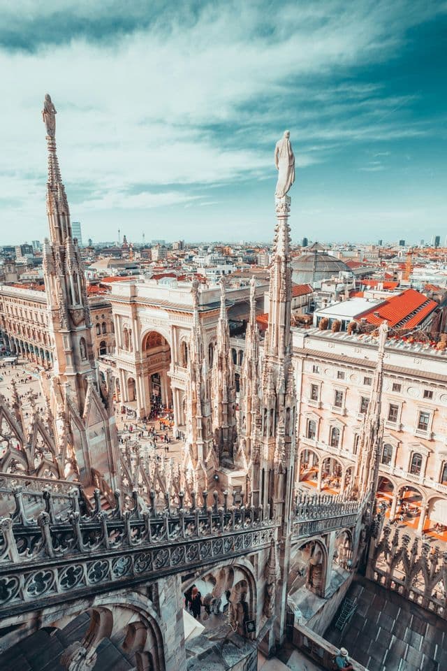 An aerial view from a Gothic cathedral's rooftop, with its intricate spires and statues overlooking a city square and buildings below.
