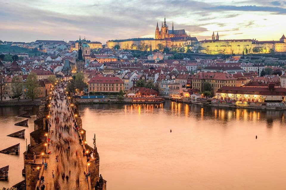 Panoramablick auf eine belebte Steinbrücke, die bei Sonnenuntergang einen Fluss überquert, mit einer beleuchteten historischen Stadt und Burg im Hintergrund.