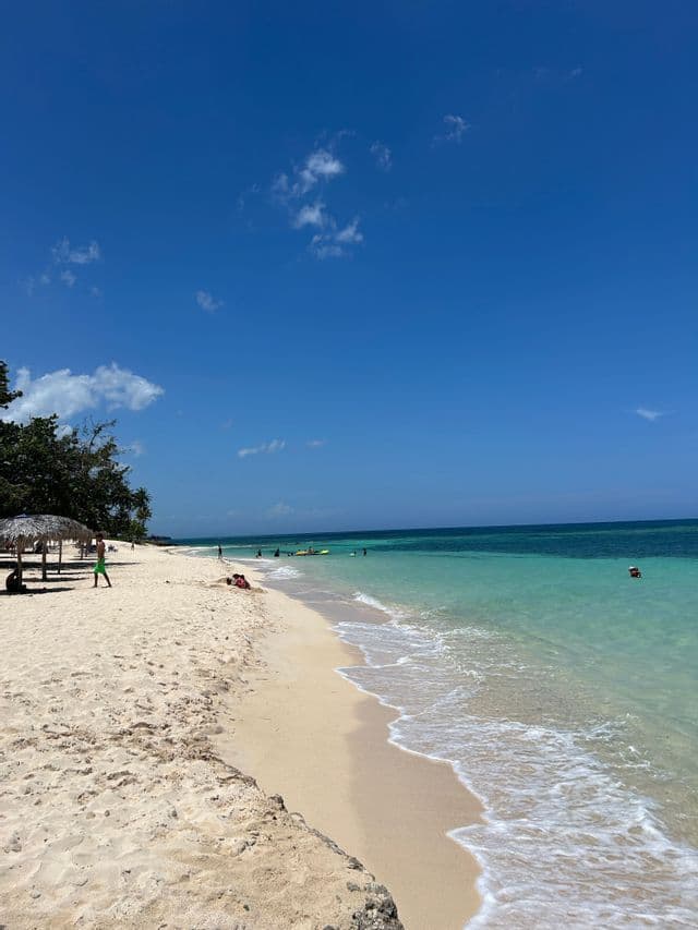 Una playa de arena blanca se encuentra con aguas cristalinas de color turquesa bajo un cielo azul brillante, con algunas personas nadando y relajándose.