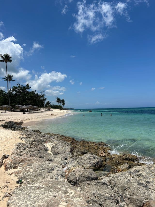 Una costa rocosa se encuentra con una playa de arena blanca con palmeras, donde la gente nada en aguas turquesas bajo un cielo azul.