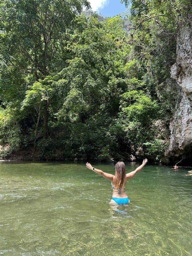 Una mujer con cabello trenzado en un bikini azul se para con los brazos extendidos en un río rodeado por un exuberante bosque verde.