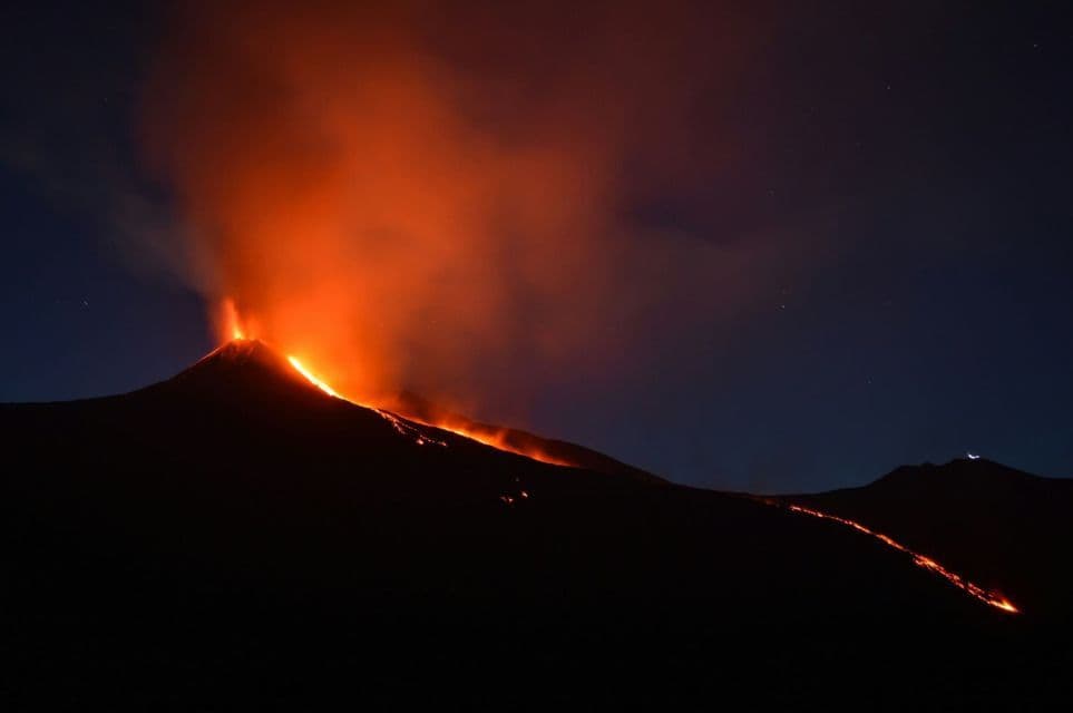 Un vulcano erutta di notte, con lava arancione incandescente che scorre lungo i suoi pendii scuri e fumo che si innalza nel cielo stellato.