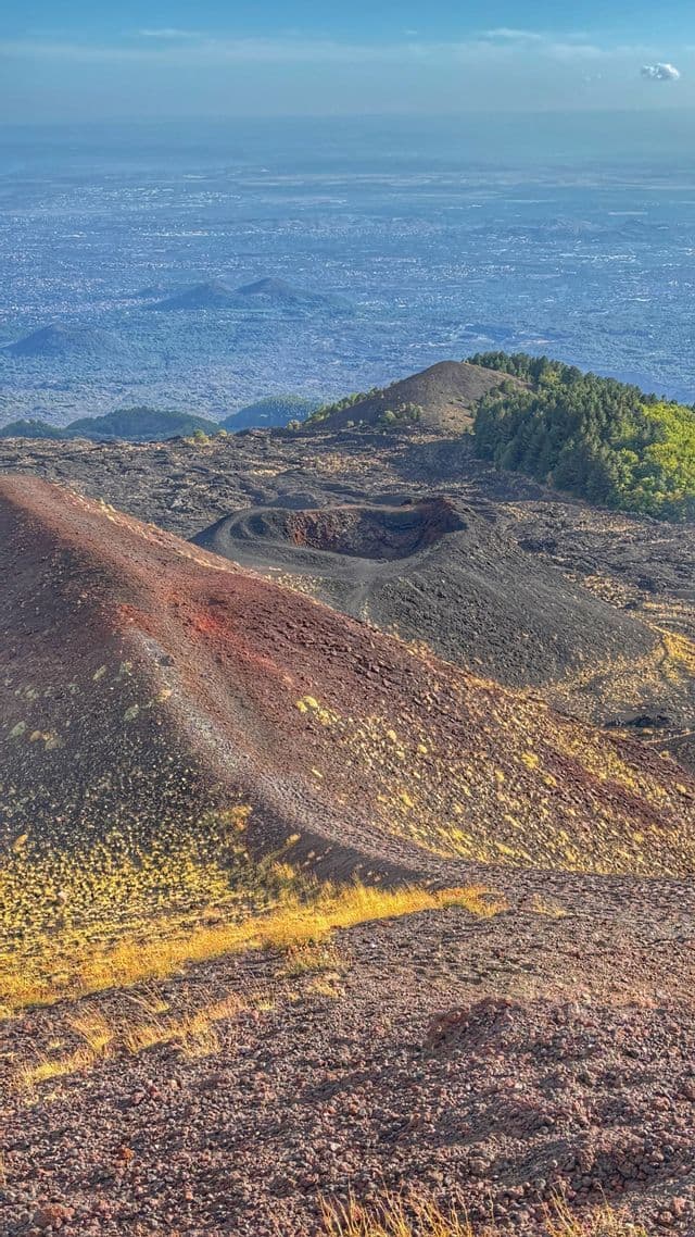 Una vista vulcanica con crateri scuri e pendii rocciosi rossastri, che si affaccia su una lontana città avvolta dalla foschia.