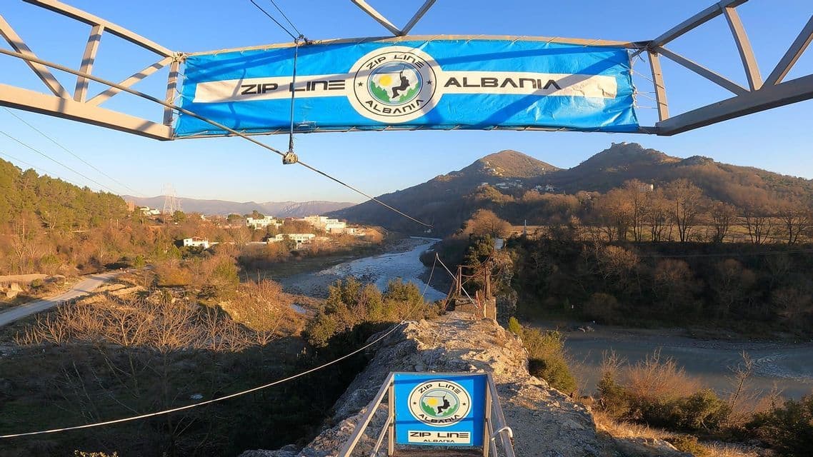 Una veduta da una piattaforma di 'Zip Line Albania' che si affaccia su una valle fluviale con montagne boscose sotto un cielo azzurro limpido.