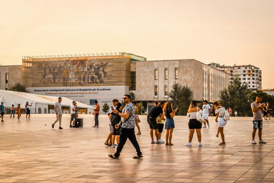 Una folla di persone passeggia e sosta in una grande piazza cittadina pavimentata al tramonto, con un edificio moderno e un grande murale sullo sfondo.