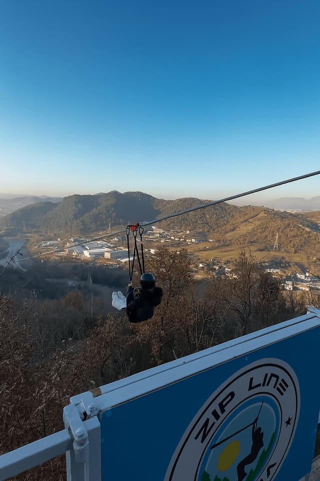 Una persona con casco e imbracatura percorre una zip line sopra una valle con colline ed edifici in una giornata serena.