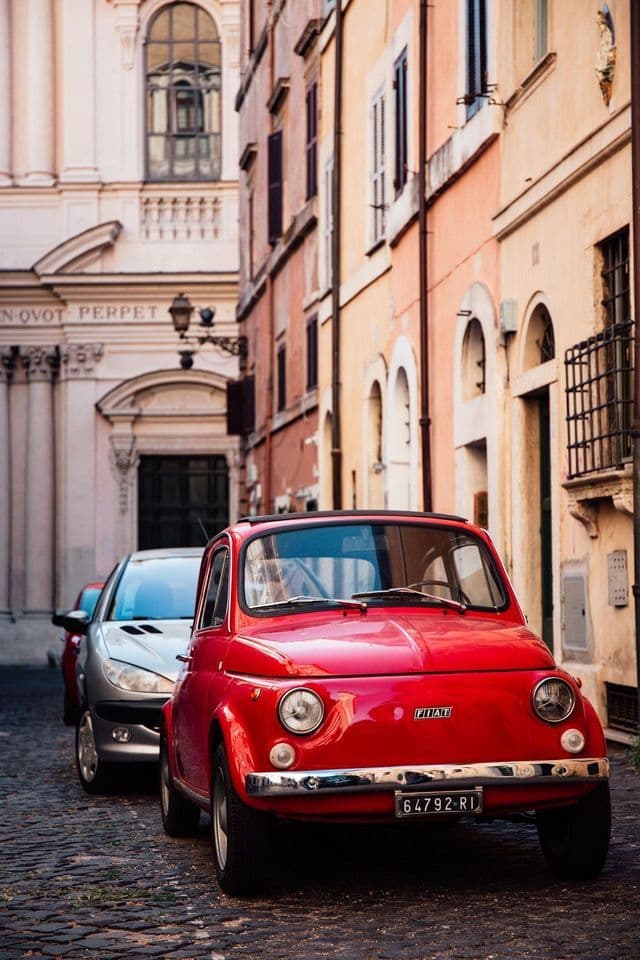 Un Fiat 500 rojo clásico estacionado en una calle estrecha de adoquines, bordeada de edificios antiguos de estilo europeo.
