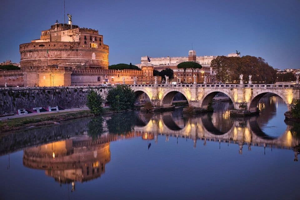 Una antigua fortaleza iluminada y un puente de piedra con arcos reflejándose en un río al atardecer.