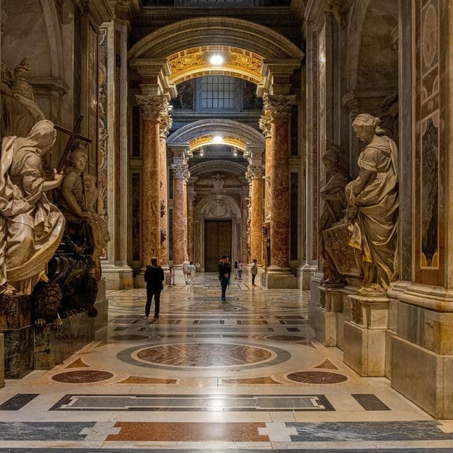 Personas caminando por un majestuoso y ornamentado salón con imponentes columnas de mármol, estatuas y un suelo estampado bajo techos abovedados.