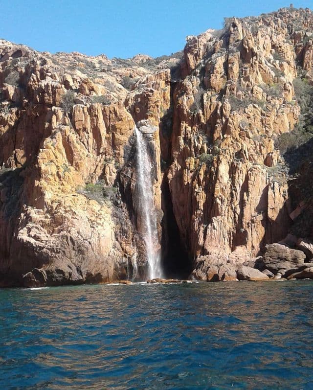 Une cascade se jette le long d'une falaise rocheuse aux teintes brun-rouge, directement dans la mer d'un bleu profond, sous un ciel dégagé.