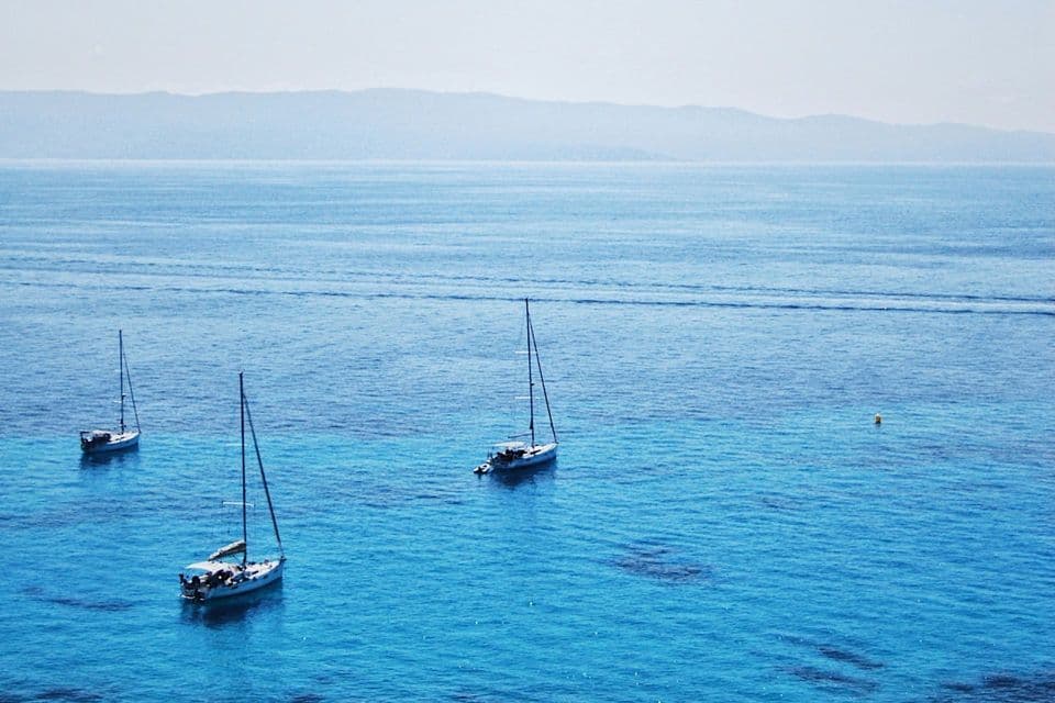 Trois voiliers flottent sur une mer calme et turquoise avec un rivage flou visible au loin.