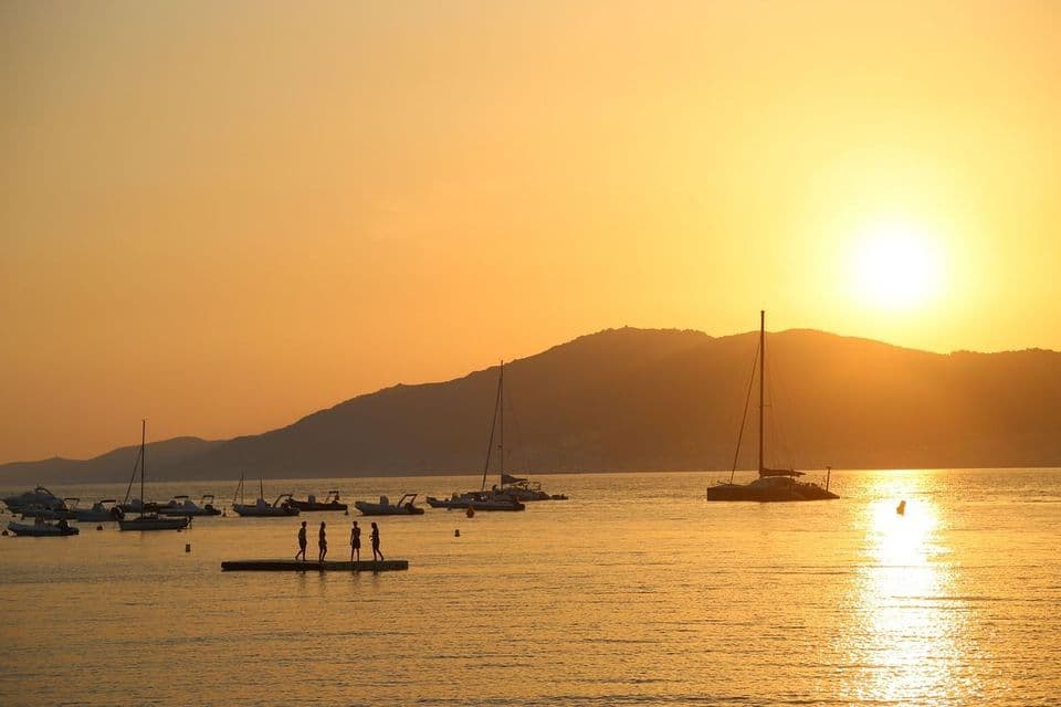 Les silhouettes de quatre personnes d'un voyage de groupe WeRoad se tiennent sur une plateforme flottante dans une baie avec des bateaux lors d'un coucher de soleil doré.