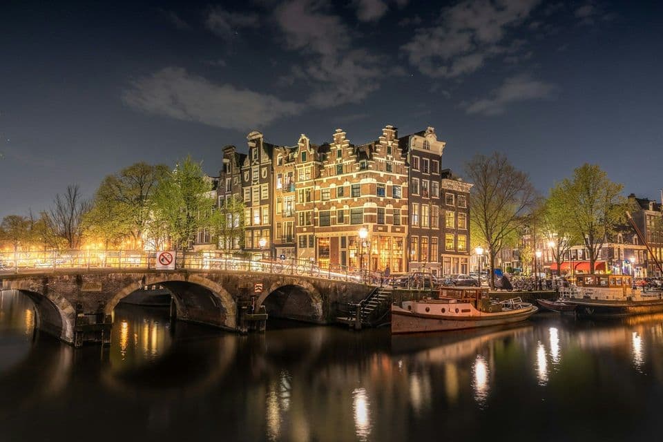 A stone bridge crosses a canal lined with illuminated gabled houses at night, their lights reflecting in the water.