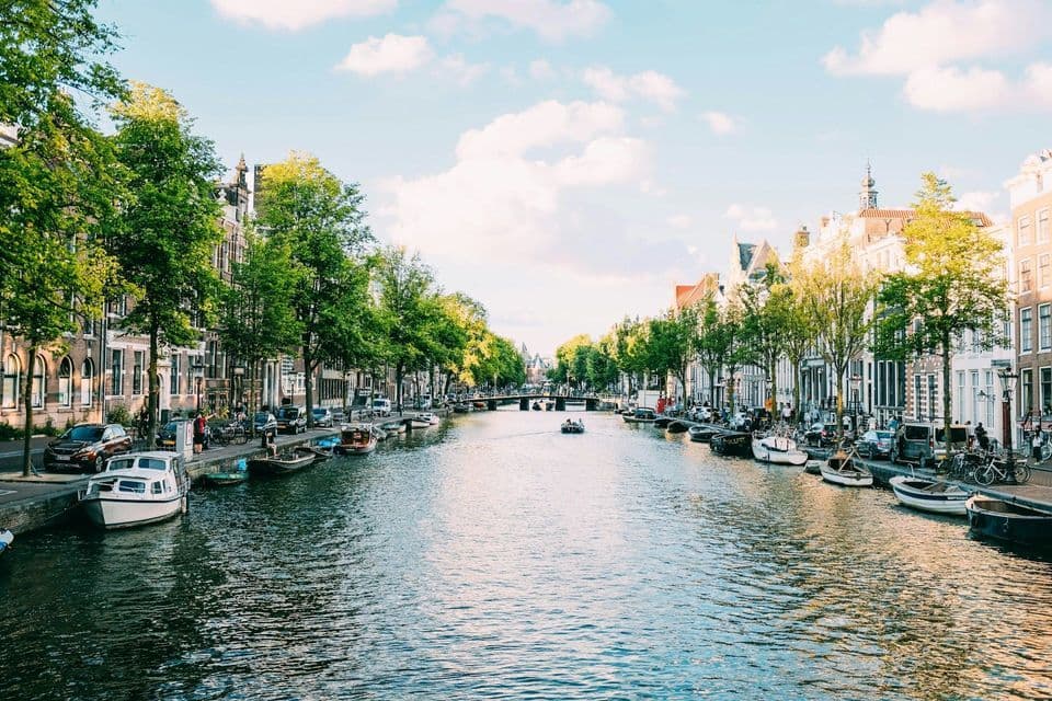 A wide city canal flows between streets lined with traditional buildings and green trees. Numerous boats are moored along the banks on a sunny day.