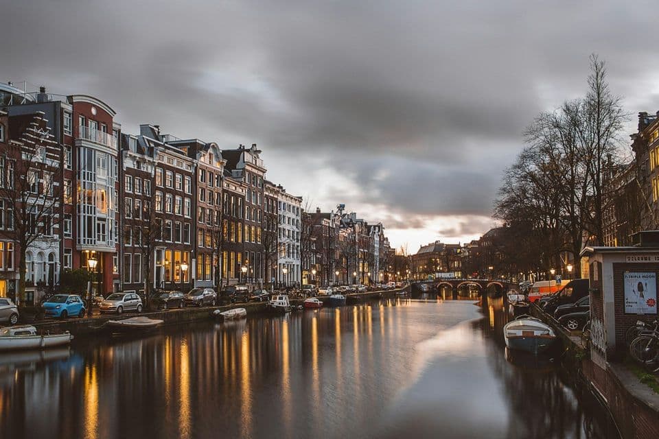 Tall buildings with lit windows line a city canal at dusk, their lights reflecting on the calm water under a cloudy sky.