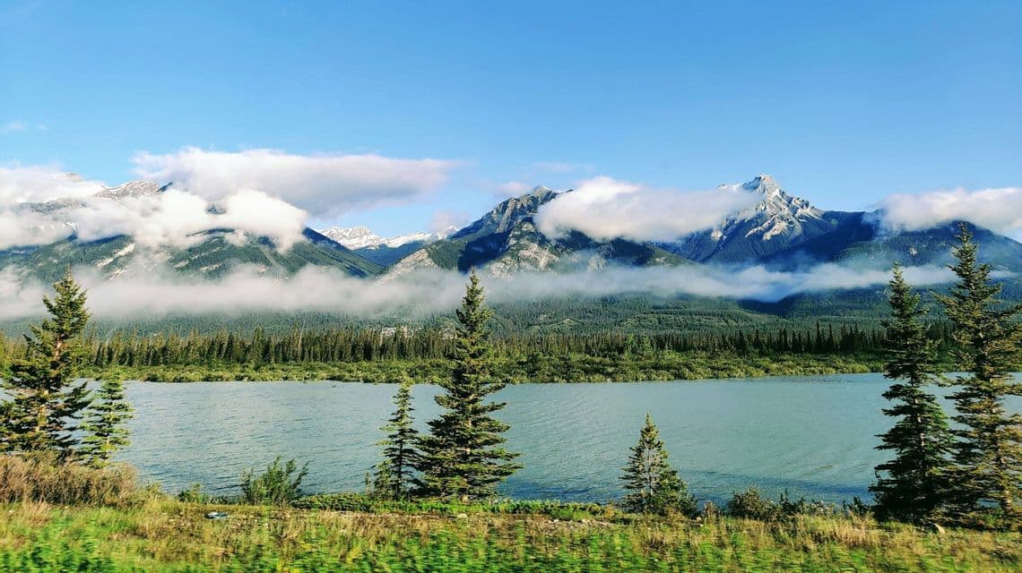 Un paesaggio con un lago sereno, una pineta e grandi montagne con le cime tra le nuvole.