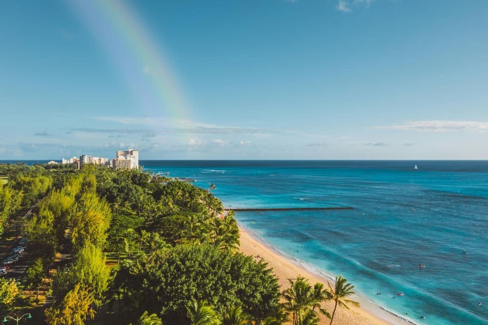 Un arcobaleno si staglia in un cielo azzurro sopra una costa tropicale con una spiaggia di sabbia, un mare turchese e alberi verdi e rigogliosi.
