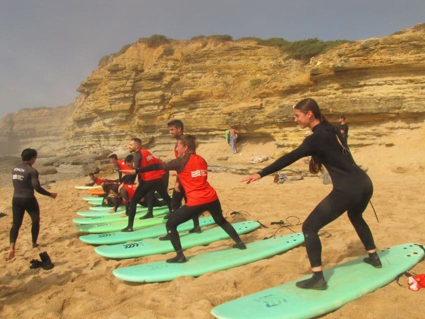 Un viaggio di gruppo WeRoad con lezione di surf su una spiaggia sabbiosa, imparando a stare in piedi sulle tavole da surf di fronte a una scogliera.