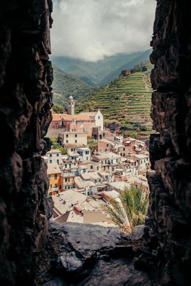 A European village with a church bell tower and terraced vineyards, framed by a dark stone opening.