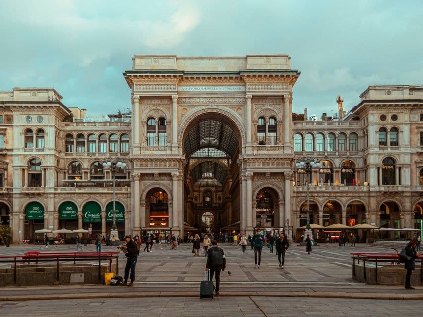 People walking across a wide, paved public square in front of a grand, historic building with a large archway.