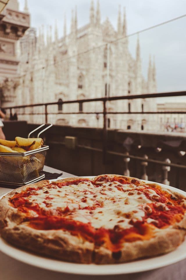 A margherita pizza and a basket of french fries on a table with a large, blurry cathedral in the background.