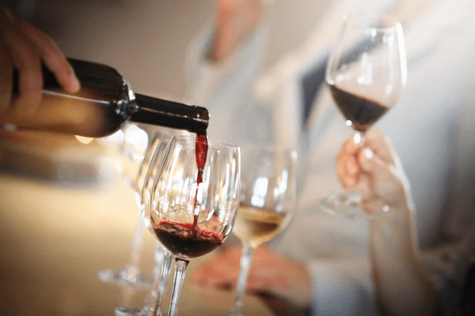 Primo piano di una mano che versa vino rosso da una bottiglia scura in un calice di vino trasparente durante una degustazione.