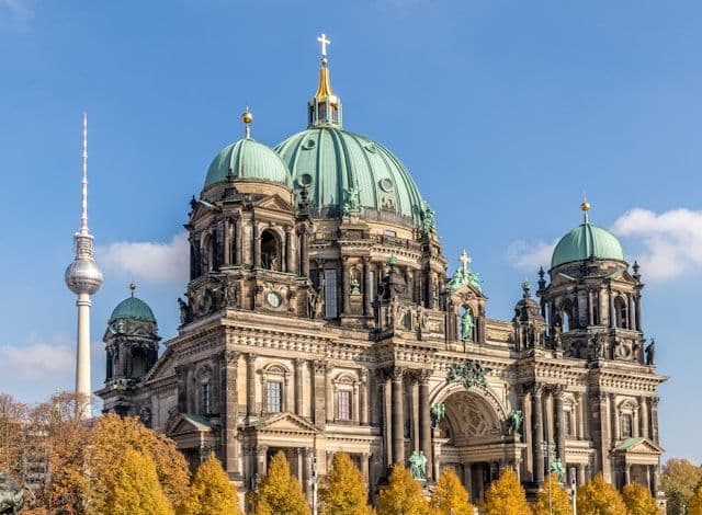 Una cattedrale sontuosa con grandi cupole verdi e una lontana torre televisiva si ergono sotto un cielo azzurro limpido sopra alberi autunnali dorati.