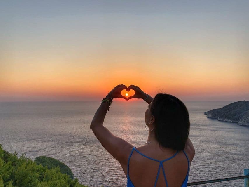 Eine Frau von hinten gesehen formt mit ihren Händen ein Herz, das die untergehende Sonne über dem Meer umrahmt.