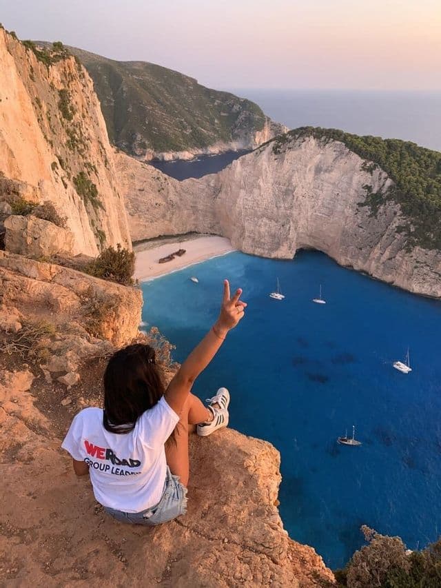 Ein WeRoad-Gruppenleiter sitzt auf einer Klippe und macht ein Friedenszeichen über einer Bucht mit strahlend blauem Wasser und Segelbooten.