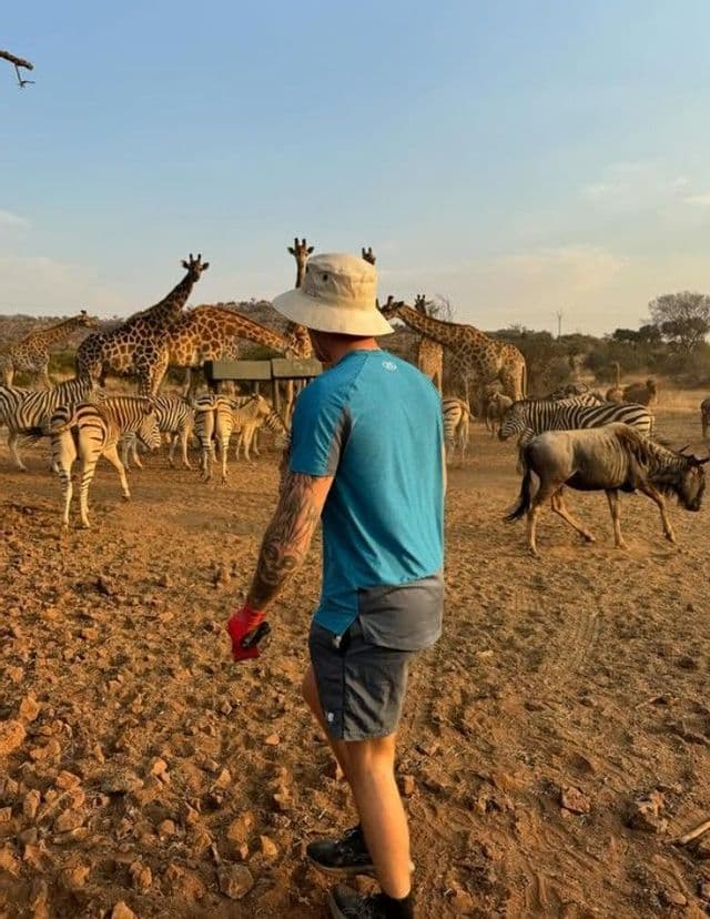Une personne portant un chapeau bob traverse un paysage sec en direction d'un troupeau de girafes, de zèbres et d'un gnou.