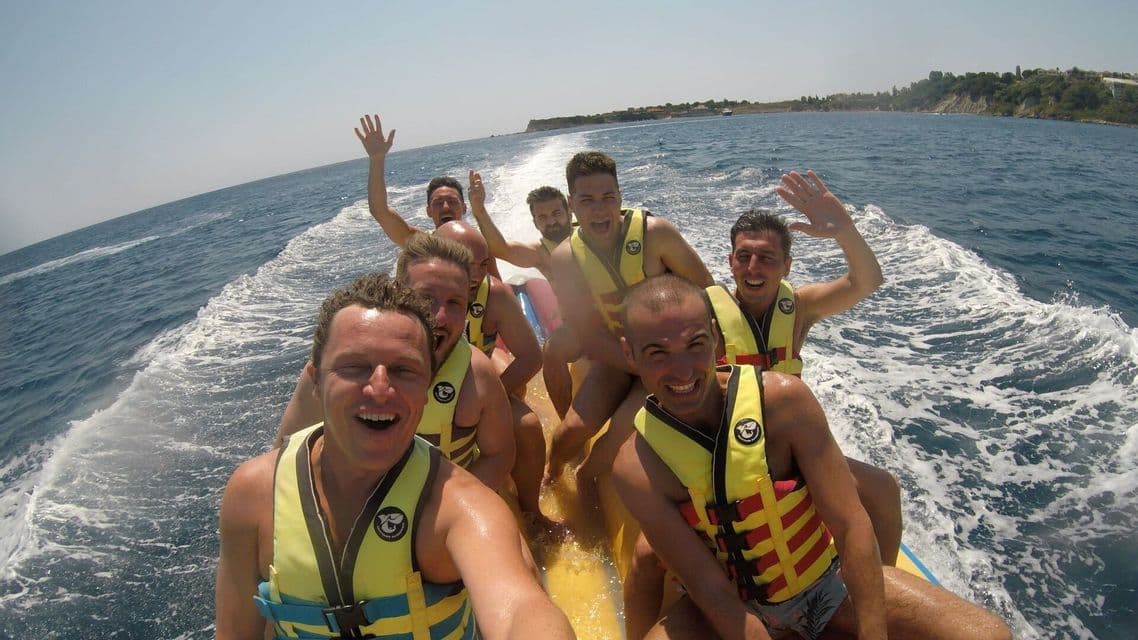 Eine WeRoad-Gruppe von Männern lacht und macht ein Selfie auf einem gelben Bananenboot im Meer.
