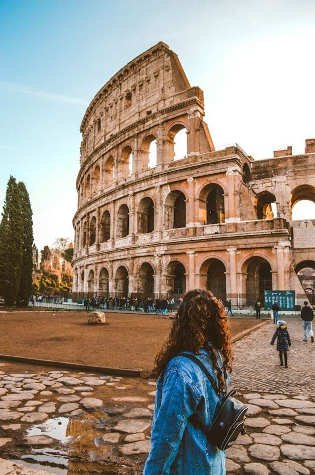 Eine Frau mit lockigem Haar, von hinten gesehen, steht auf einem Kopfsteinpflasterplatz und blickt auf ein großes, antikes steinernes Amphitheater.
