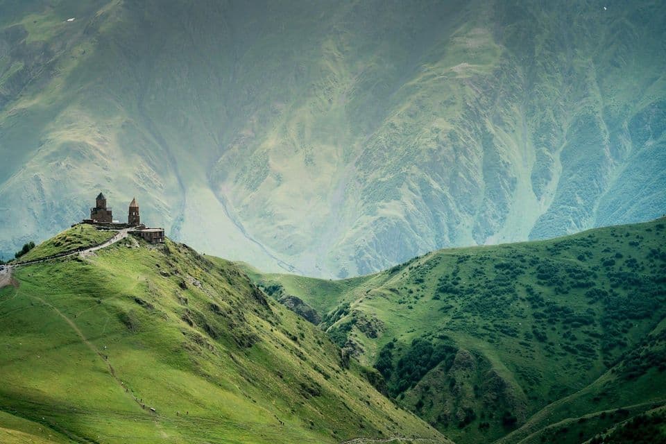 An old stone church stands on a green hill, with massive, misty mountains rising in the background.