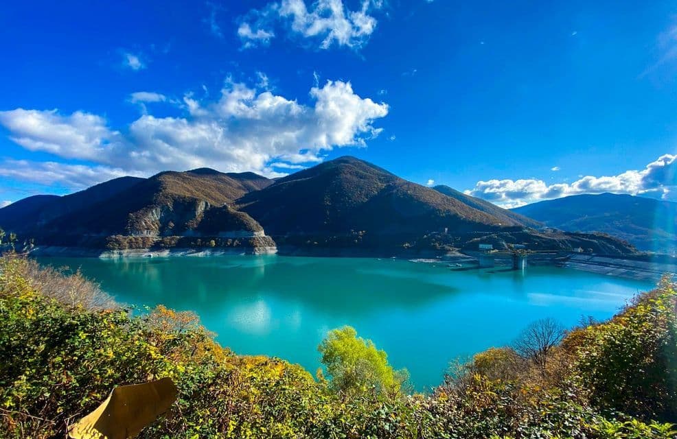A turquoise lake surrounded by mountains with autumn-colored trees under a bright blue sky with scattered clouds.