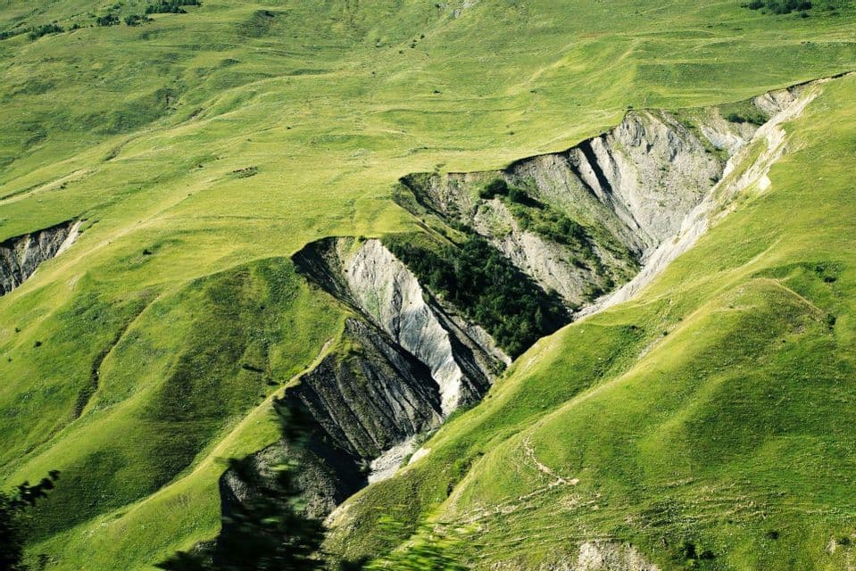 Sunlight illuminates rolling green hills with a deep ravine that exposes layers of grey and dark stratified rock.