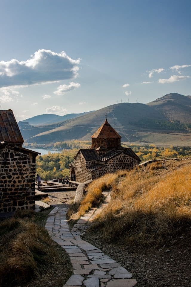 A winding cobblestone path leads up a grassy hill to an ancient stone church, with a lake and mountains in the distance.
