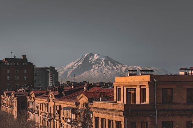 A view over warm-toned city rooftops reveals a large, snow-capped mountain rising in the distance under a hazy sky.
