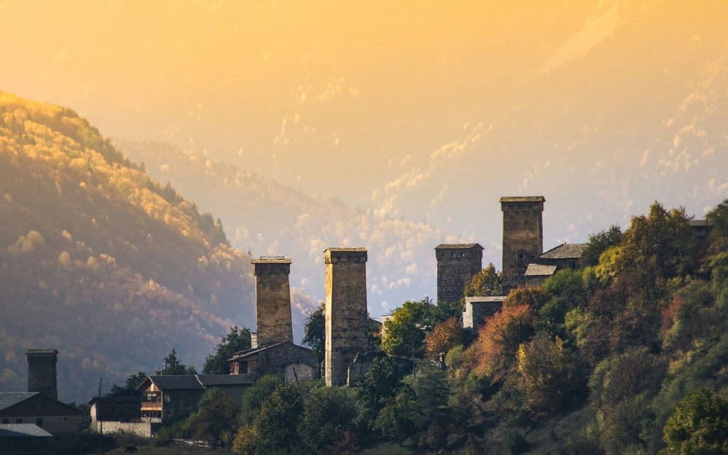 Several stone towers stand in a village on a forested mountain during a hazy, golden sunset.