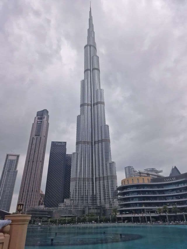 Eine Untersicht des Wolkenkratzers Burj Khalifa, der einen bewölkten Himmel durchstößt, mit umgebender moderner Architektur und einem türkisfarbenen Pool.