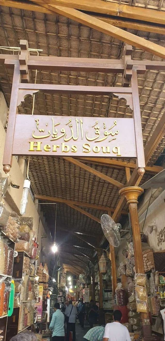 Ein Holzschild mit der Aufschrift 'Herbs Souq' hängt über einer belebten, schmalen Gasse in einem traditionellen, überdachten Markt mit Ständen auf beiden Seiten.