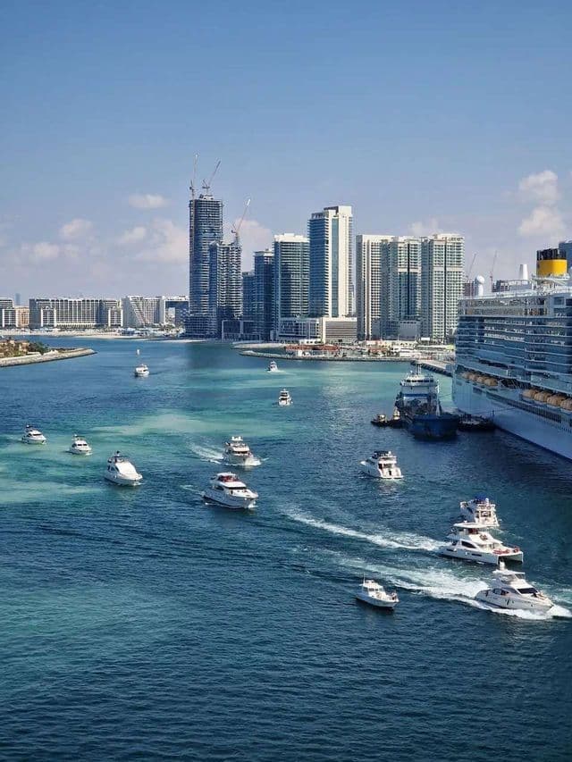 Zahlreiche weiße Yachten segeln auf türkisfarbenem Wasser in einem Hafen neben einem großen Kreuzfahrtschiff, mit einer modernen Stadtsilhouette im Hintergrund.
