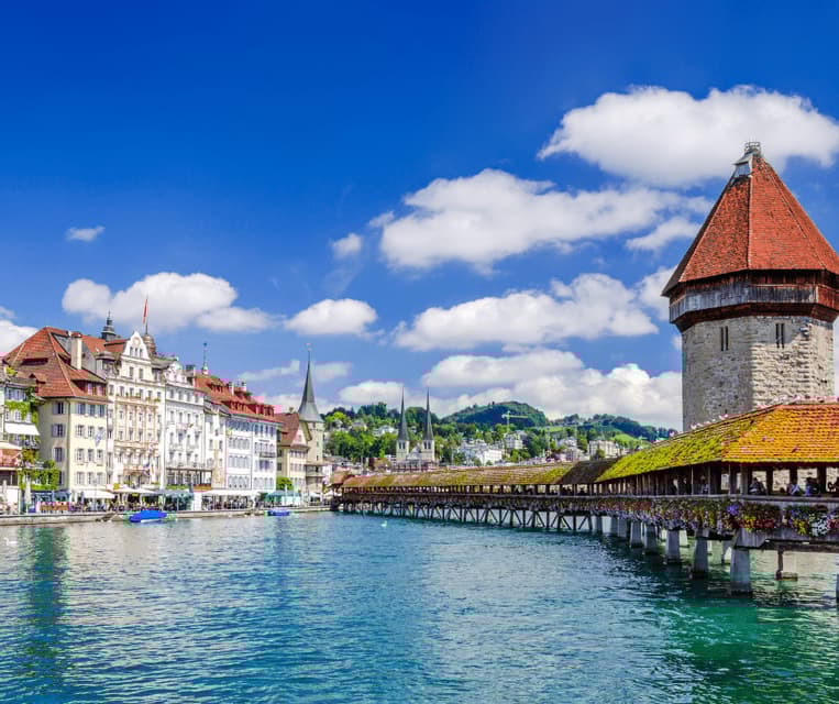 A covered wooden bridge with a stone tower spans a turquoise river next to a city waterfront under a blue sky.