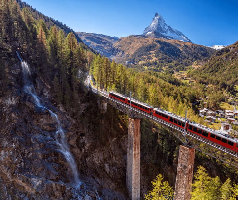 A red train crosses a high bridge over a mountain valley, with a waterfall on the left and a snow-capped peak in the background.