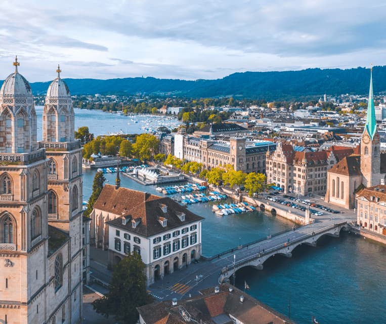 Aerial view of a historic city with twin church towers overlooking a river with a bridge and docked boats, with hills in the background.